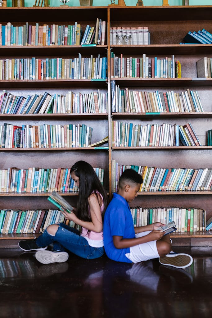 Two children sitting back to back reading books in a library setting.